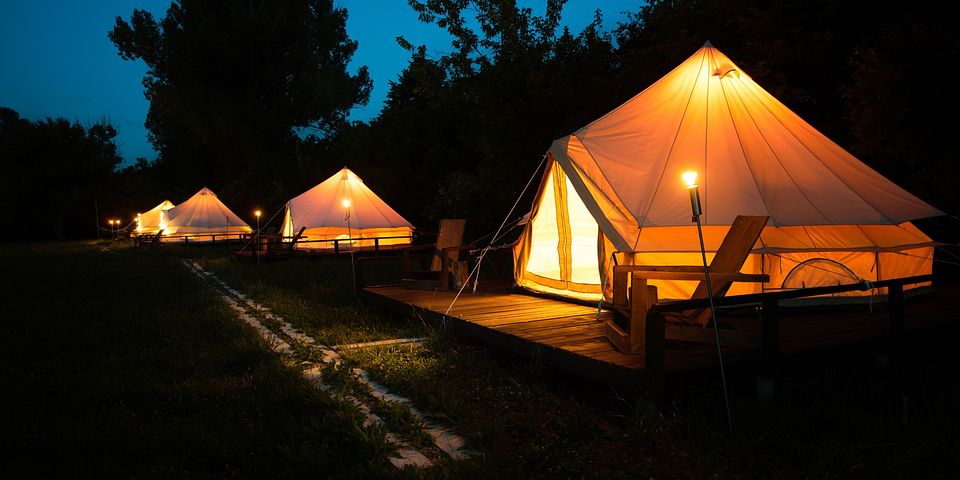 Glamping tents with warm lights at night, set on wooden platforms amidst a lush forest setting.