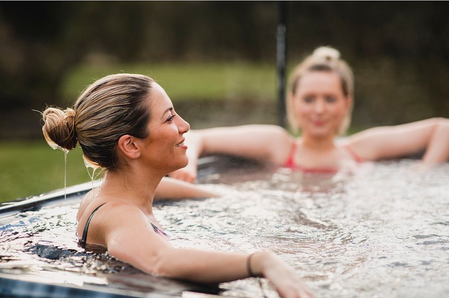 Two women relaxing in a hot tub outdoors with serene expressions, surrounded by a natural landscape.