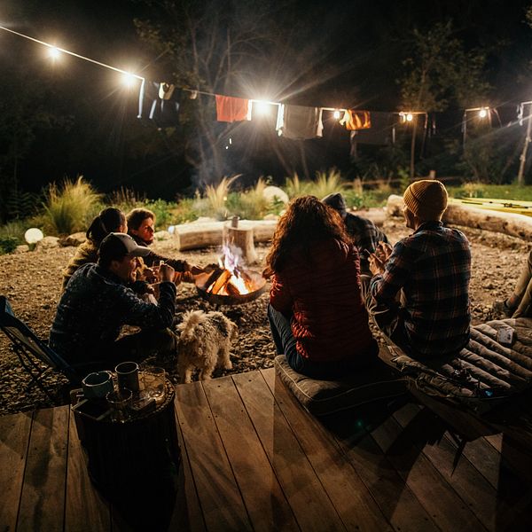 A group of people sit around a campfire with lights above it at night and enjoy each other's company in the outdoors.